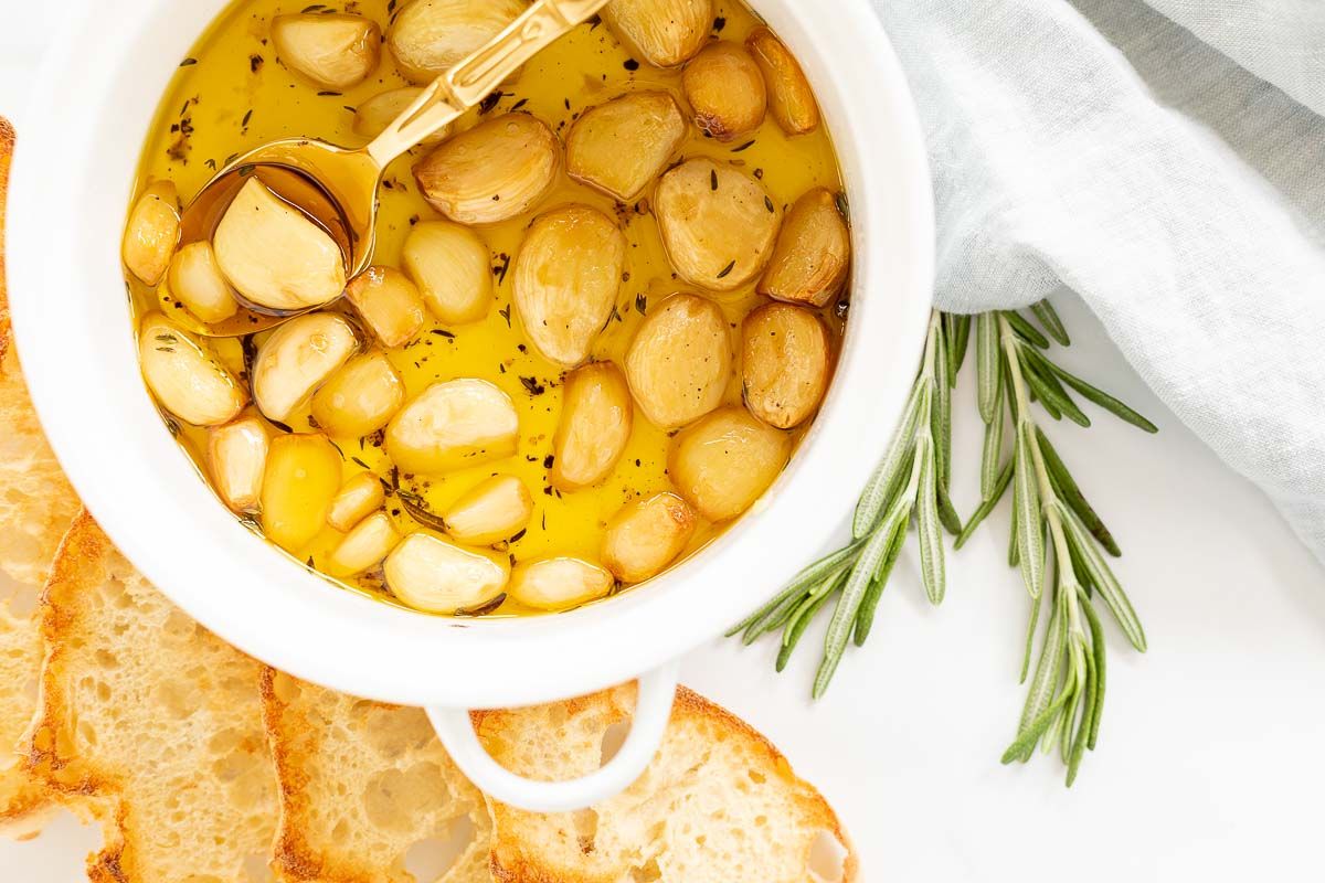 A white bowl full of garlic confit, surrounded by crostini and fresh rosemary.