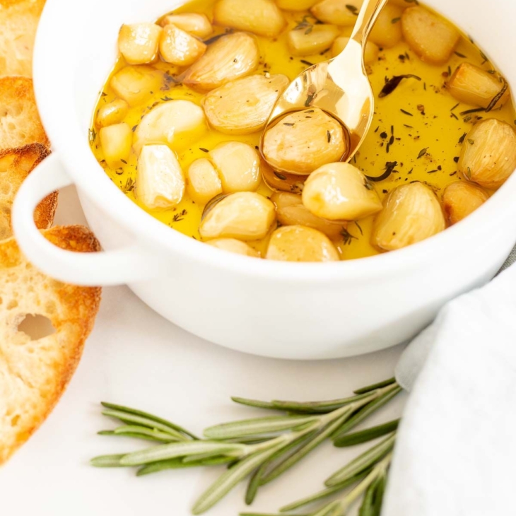 A white bowl full of garlic confit, surrounded by crostini and fresh rosemary.
