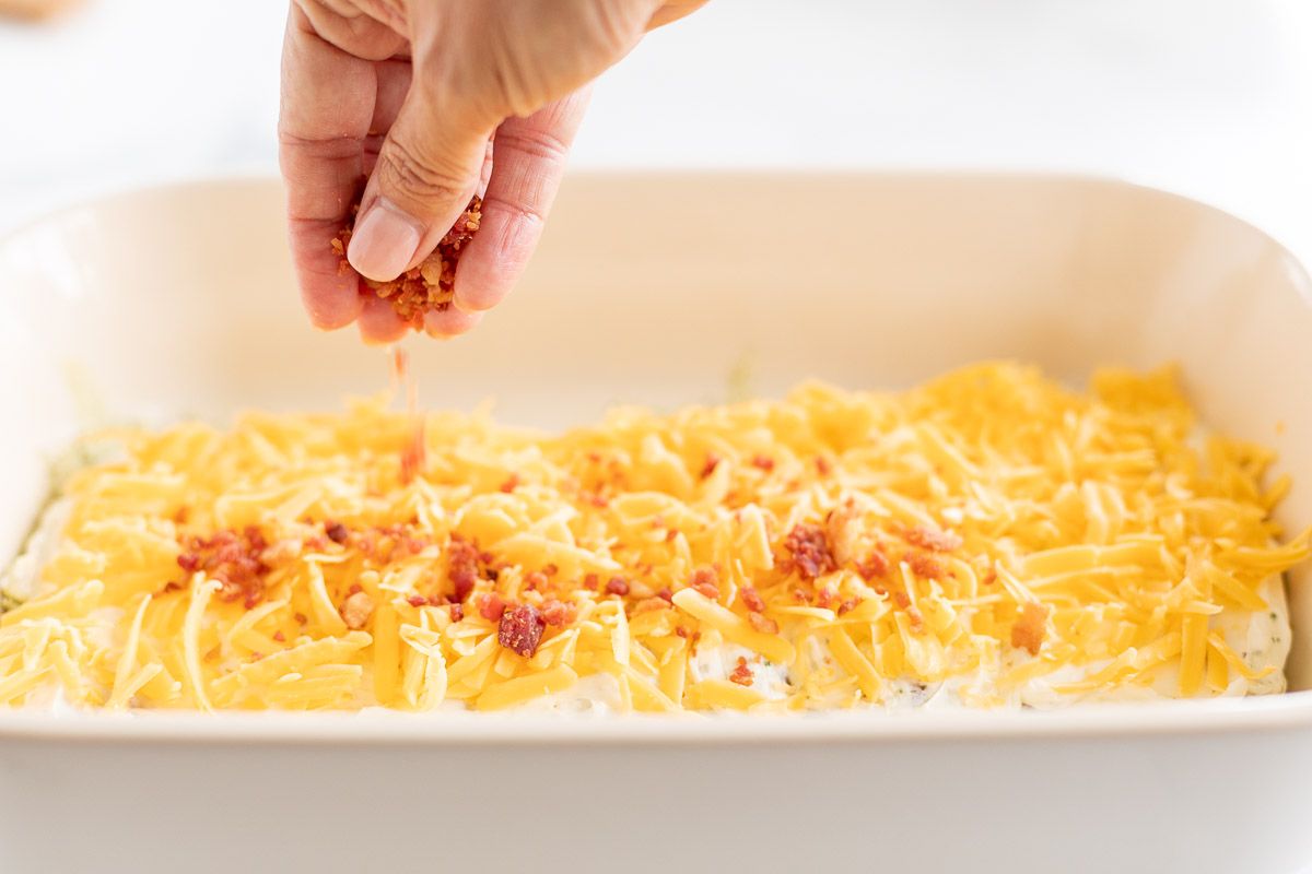 A hand spreading chopped bacon over a white baking dish full of crack chicken, prior to baking.