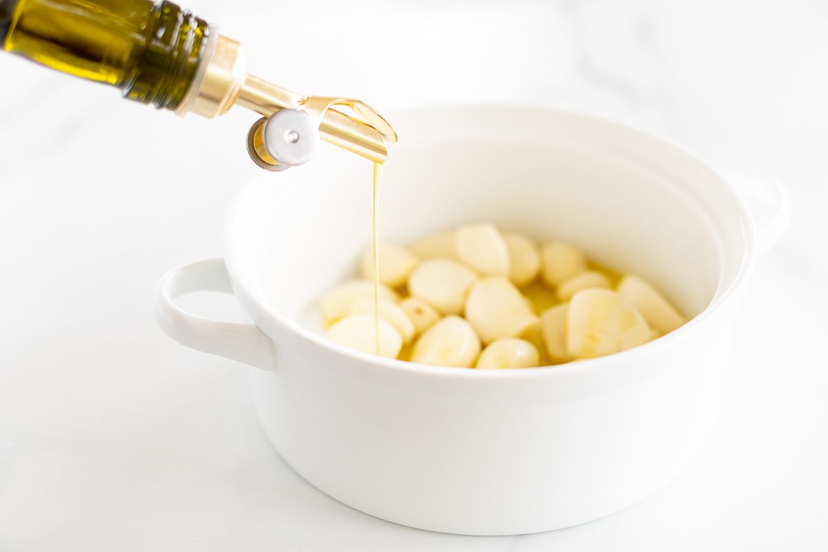 A white bowl of garlic cloves, and a bottle of olive oil pouring over the top