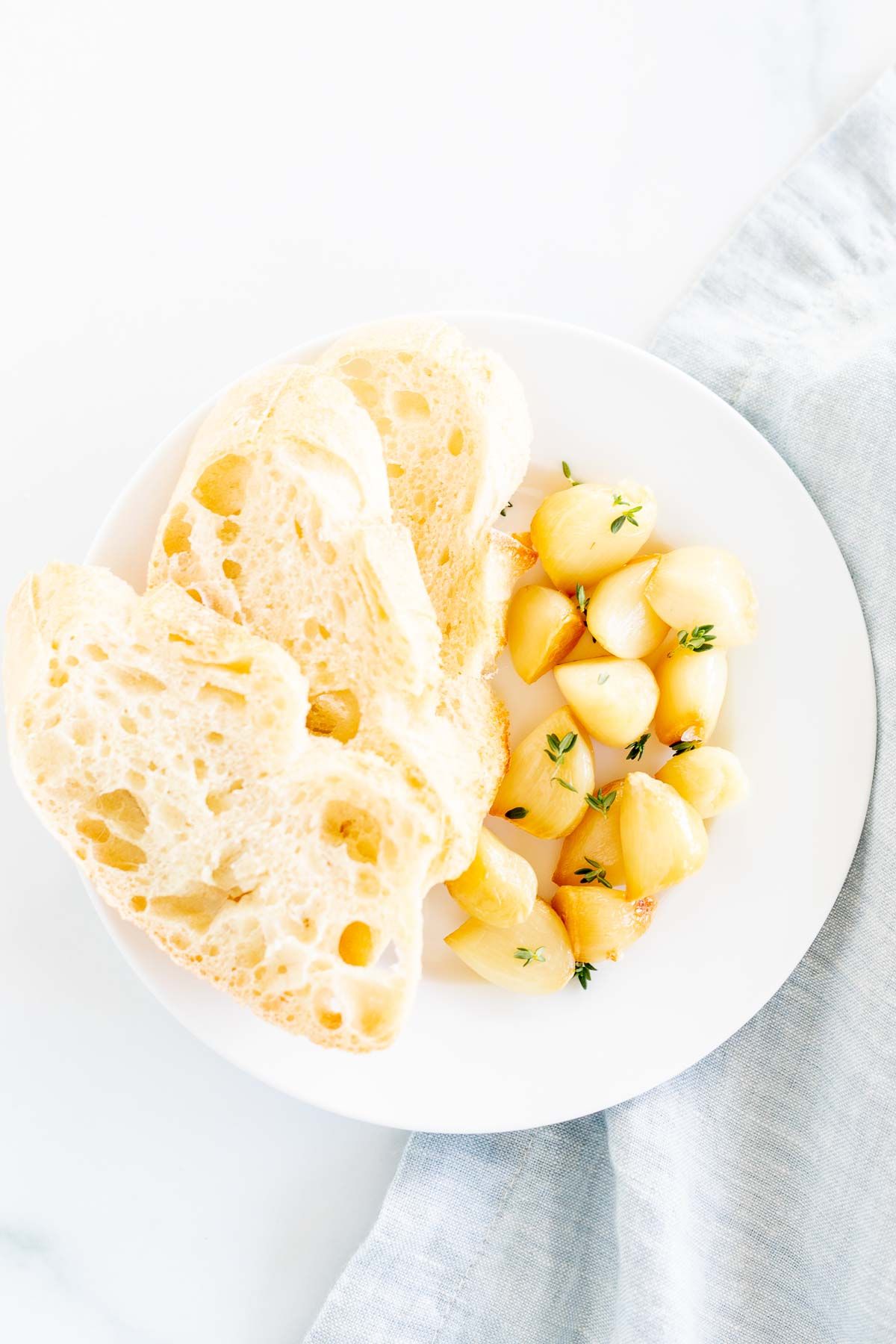 A white plate with sliced French bread and cloves of roasted garlic TeamJiX