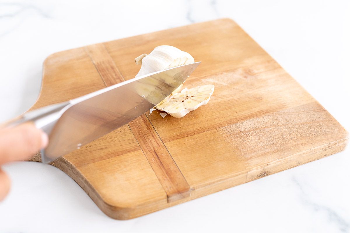 A knife slicing the ends off of a garlic bulb on a wooden cutting board TeamJiX