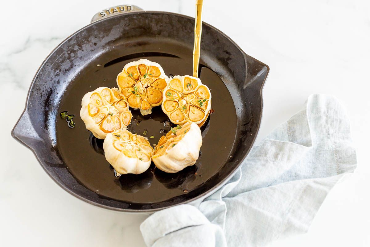 A gold spoon lifting up a head of roasted garlic from a cast iron pan