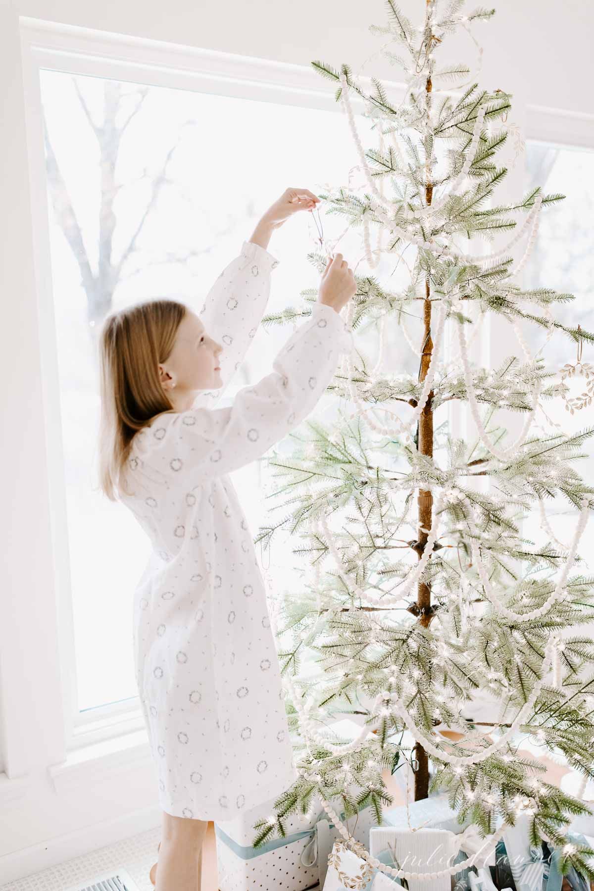 A little girl in a Christmas night shirt decorating a sparse Christmas tree, with blue packages below.