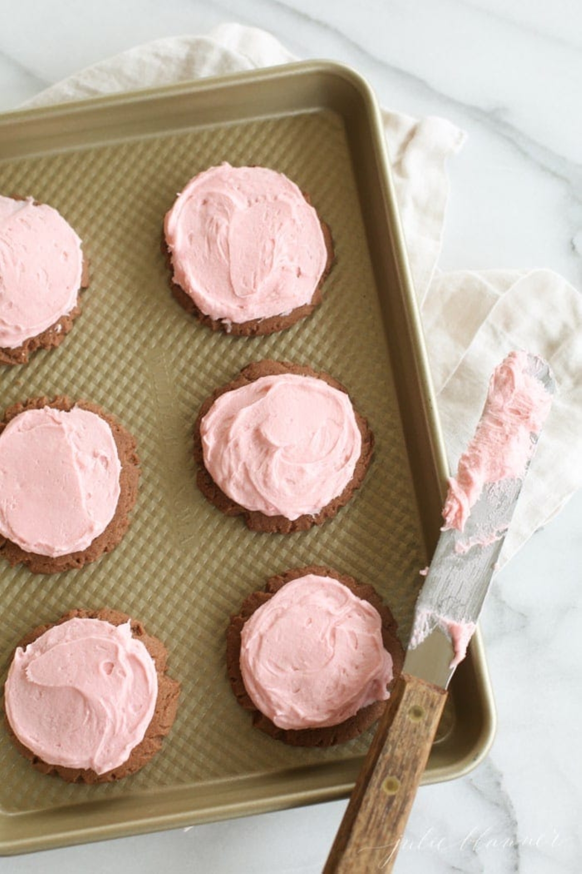 Chocolate sugar cookies iced with pink buttercream icing, on a gold cookie sheet.