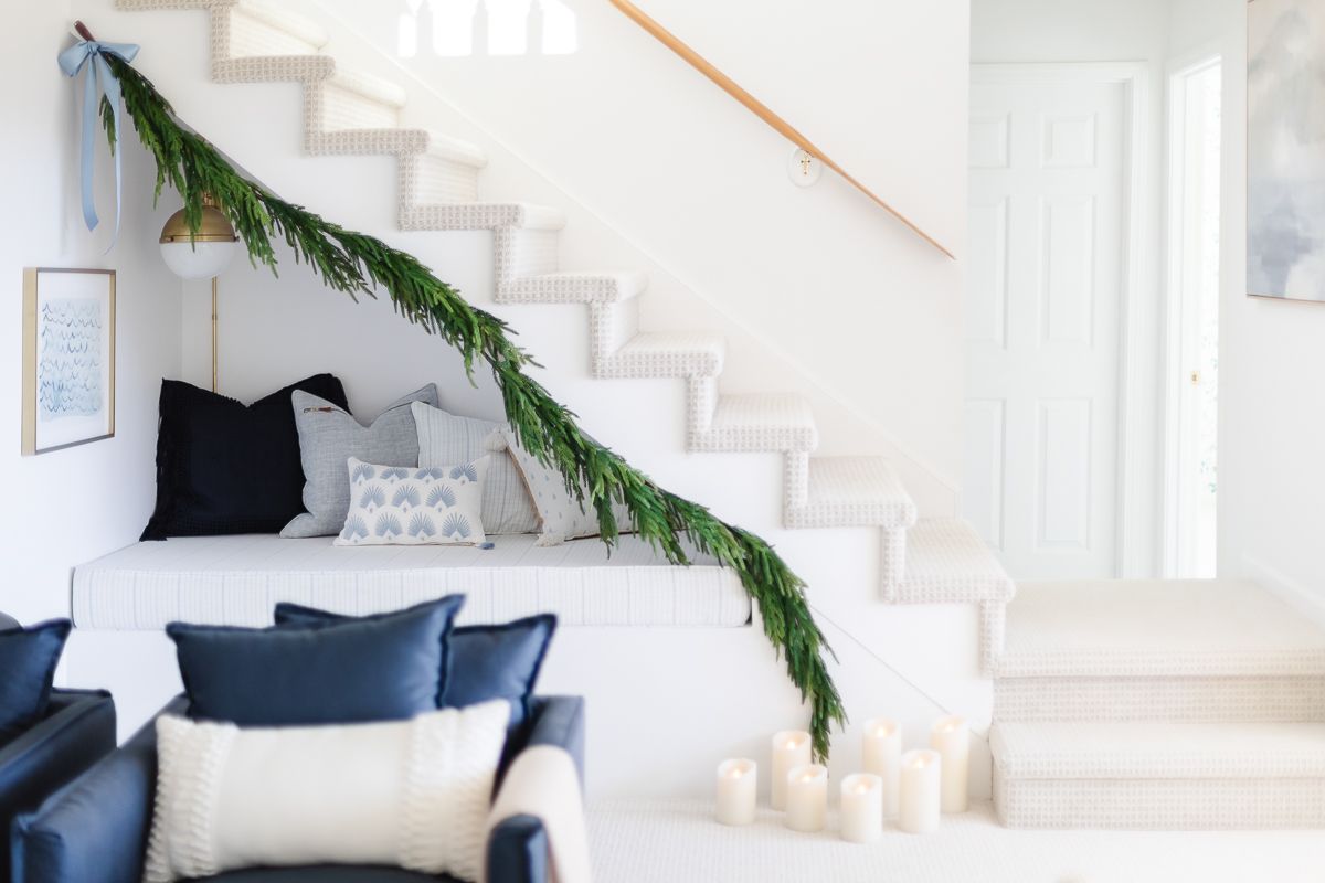 A navy and white reading nook under a staircase in a coastal Christmas living room, garland hangs above.