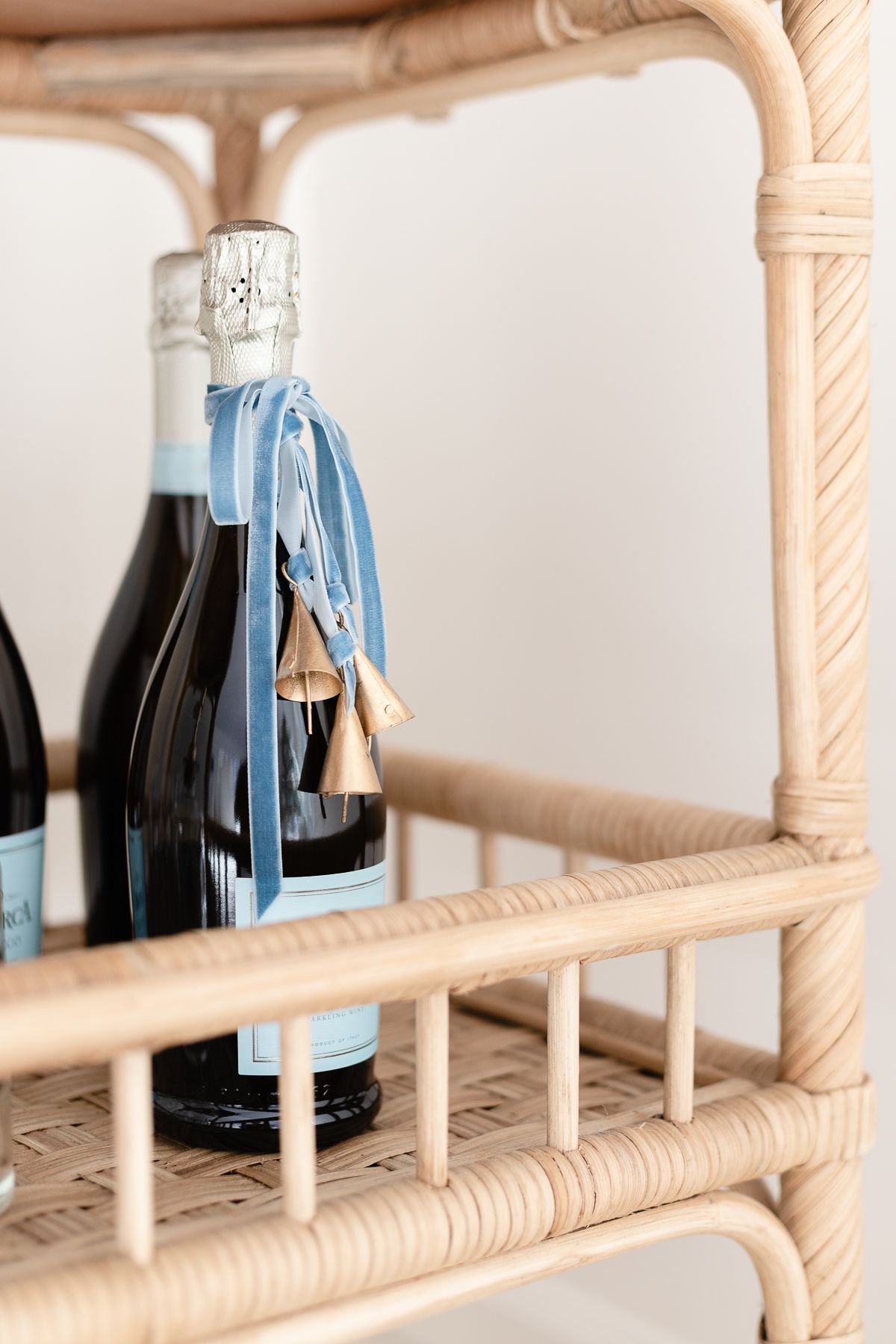 A bottle of champagne tied with a velvet bow and brass bells on a rattan bar cart.