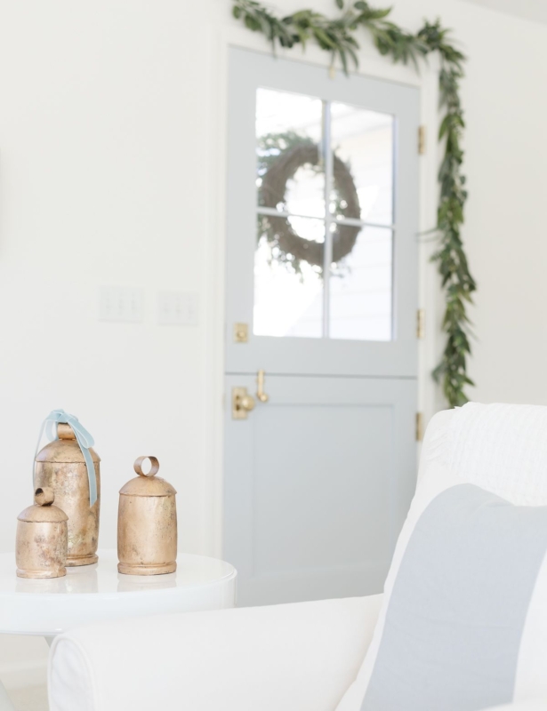 A blue Dutch door accented with a greenery garland, and brass bells on a white table in a coastal Christmas living room.