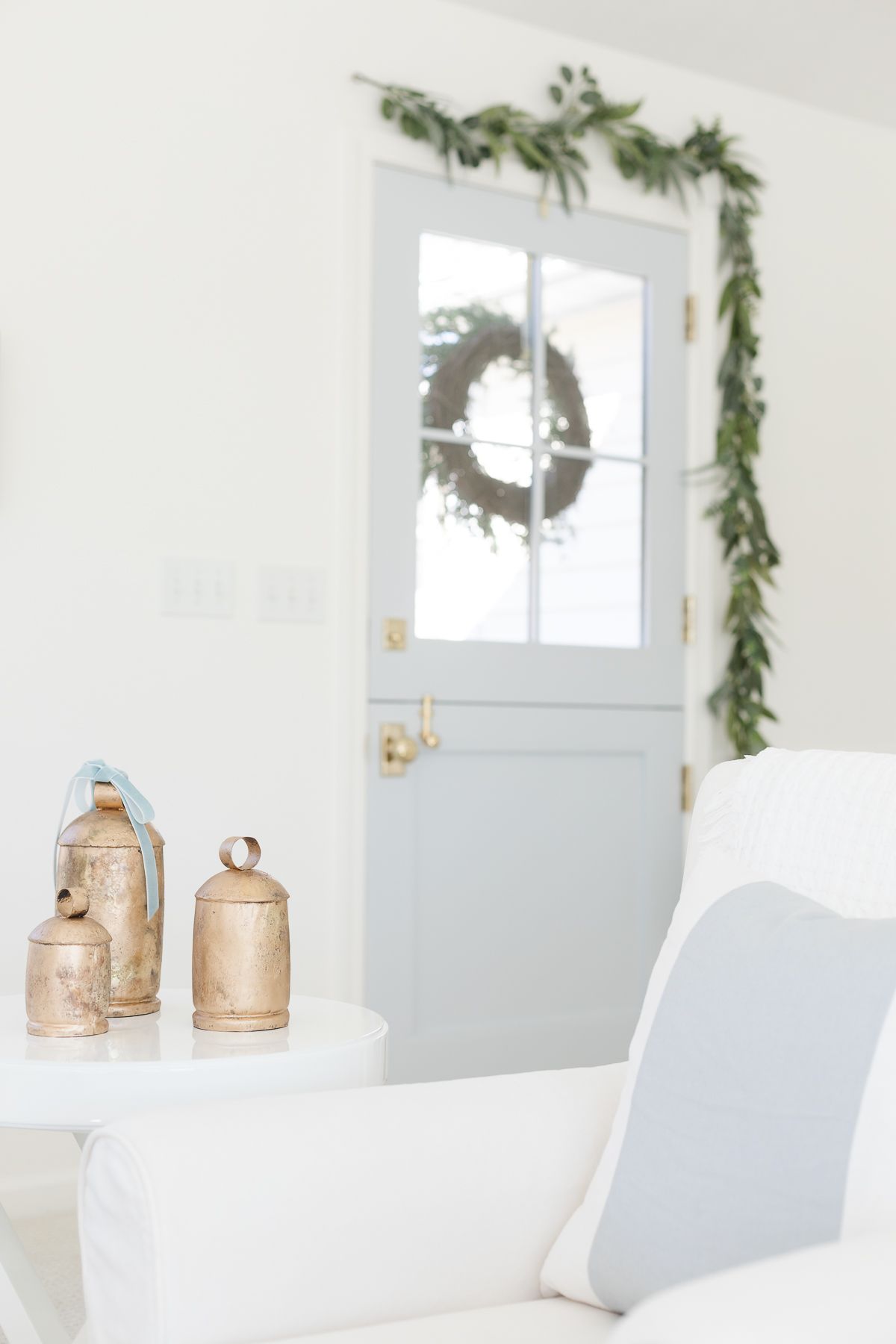A blue Dutch door accented with a greenery garland, and brass bells on a white table in a coastal Christmas living room.