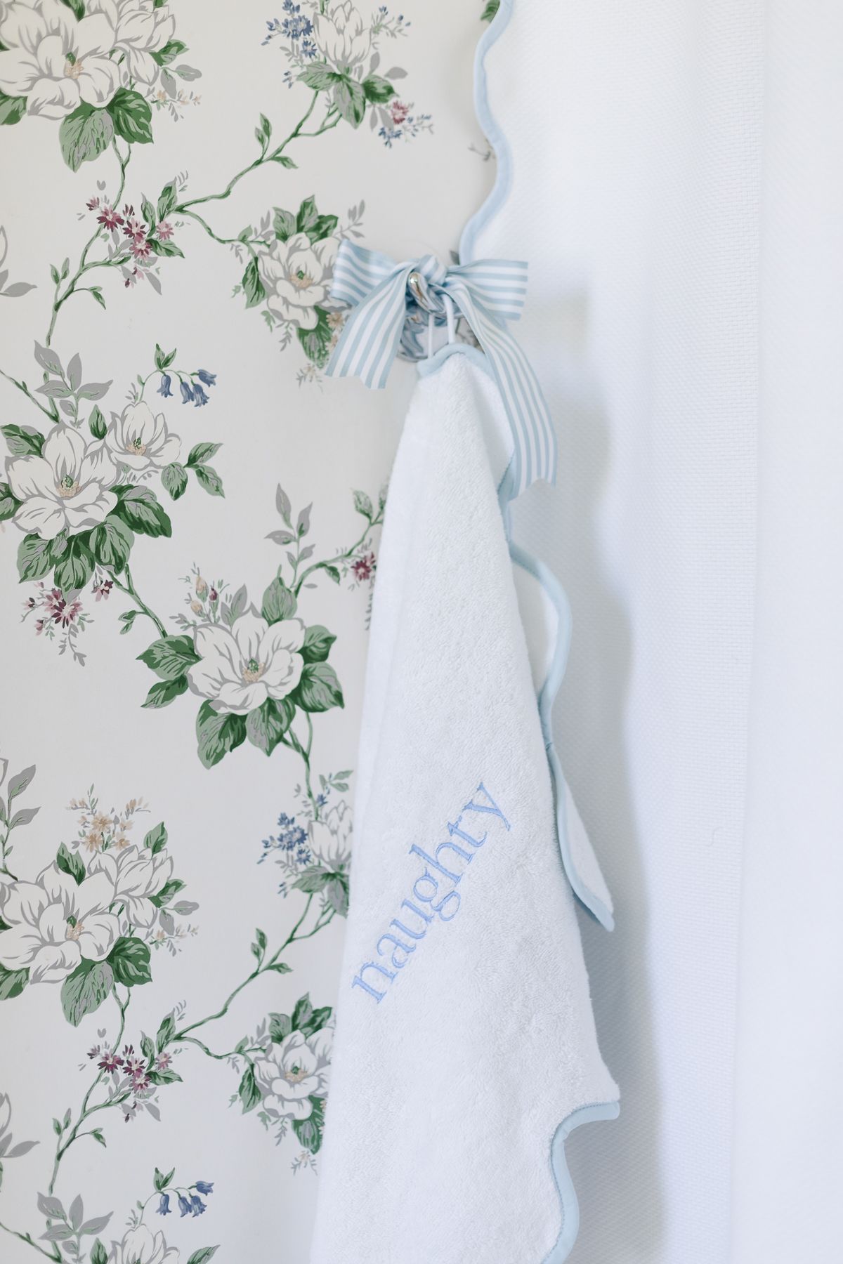 A blue and white towel that is embroidered with the word "naughty" in a floral wallpapered bathroom.