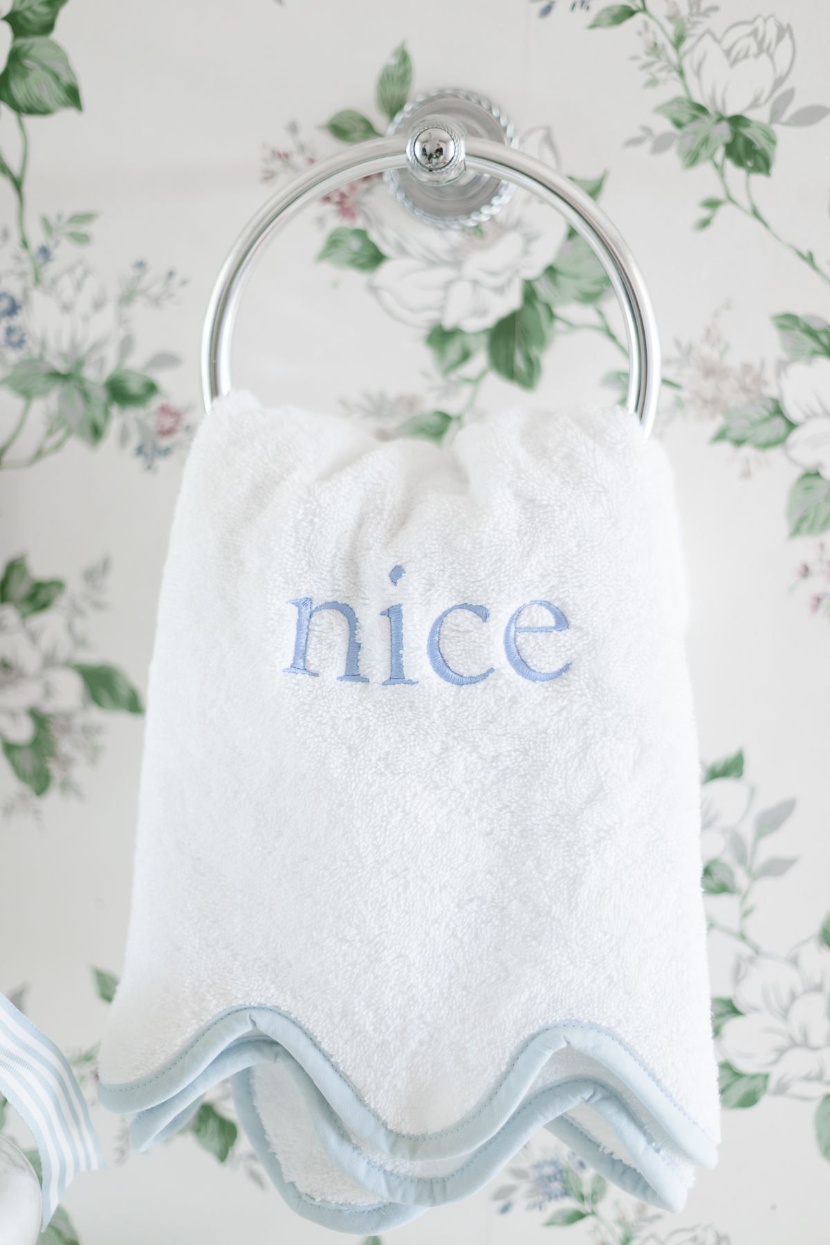 A blue and white towel that is embroidered with the word "nice" in a floral wallpapered bathroom.