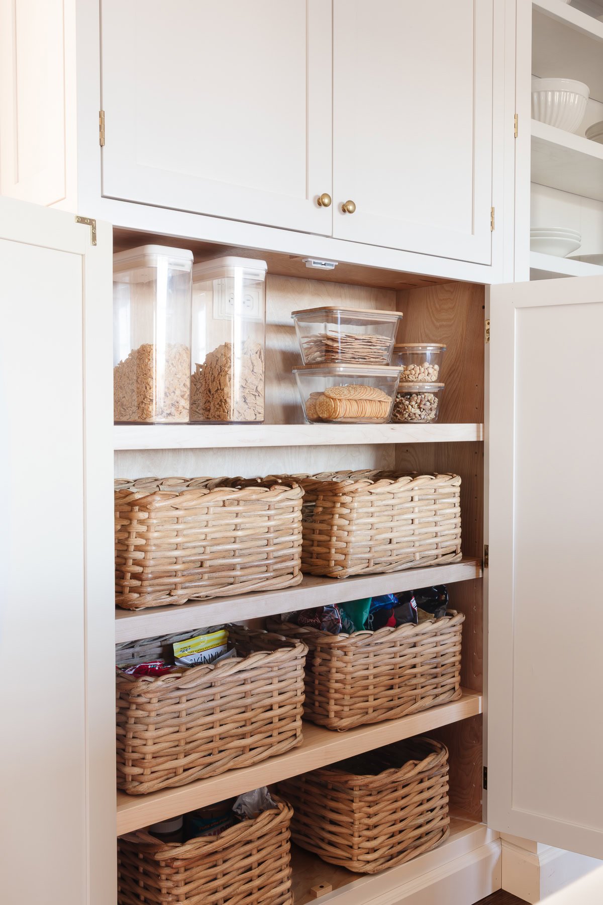 A decluttered pantry cabinet with open doors reveals organized shelves, clear containers of dry goods, and multiple wicker baskets storing various items.