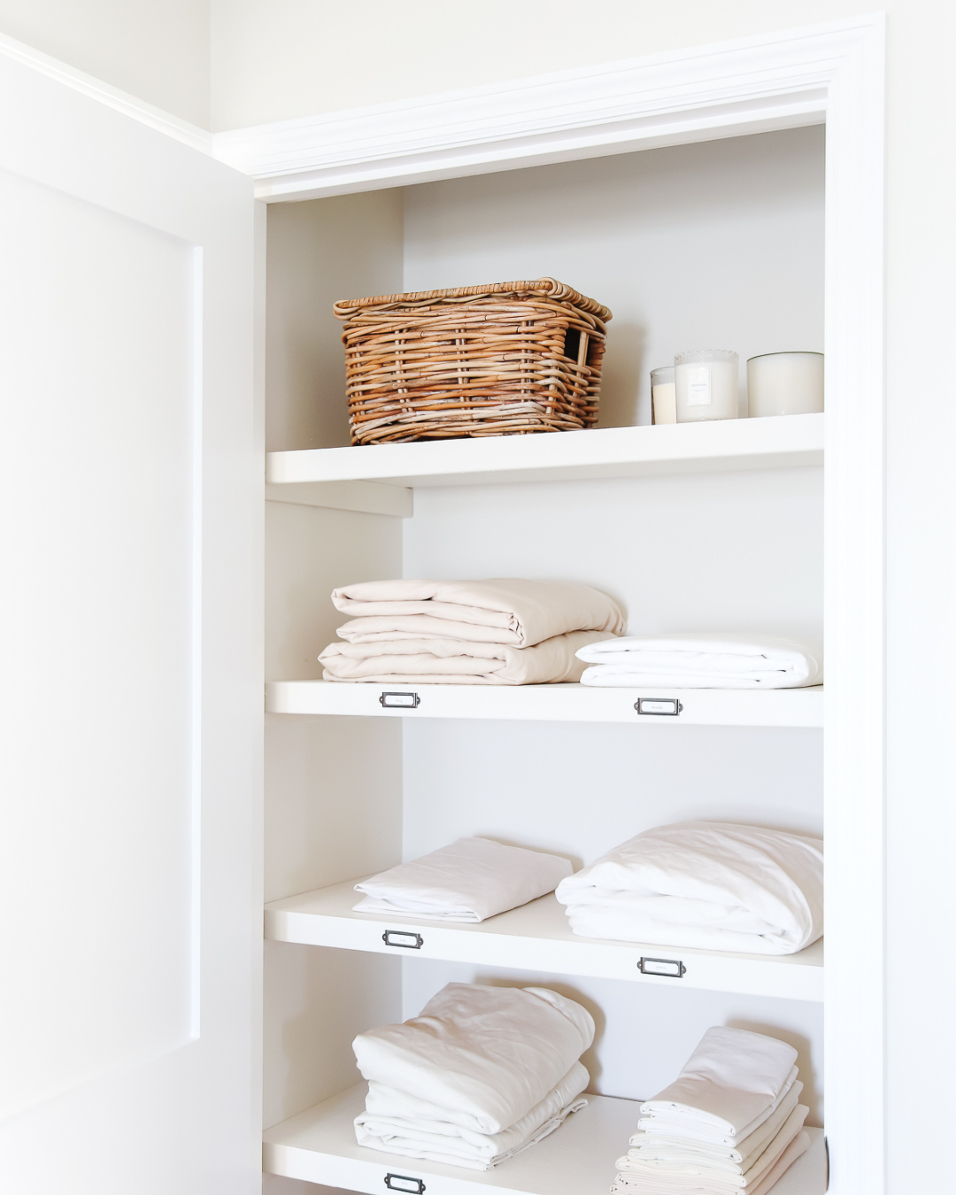 A neatly organized, decluttered linen closet features folded white sheets on shelves, a wicker basket, and three candles on the top shelf.