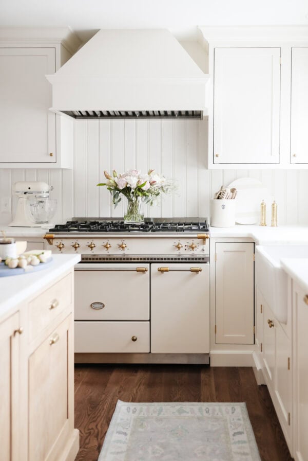 A white kitchen with a vintage-style stove, gold hardware, and decluttered white cabinets features wood floors and a vase of flowers on the stove.
