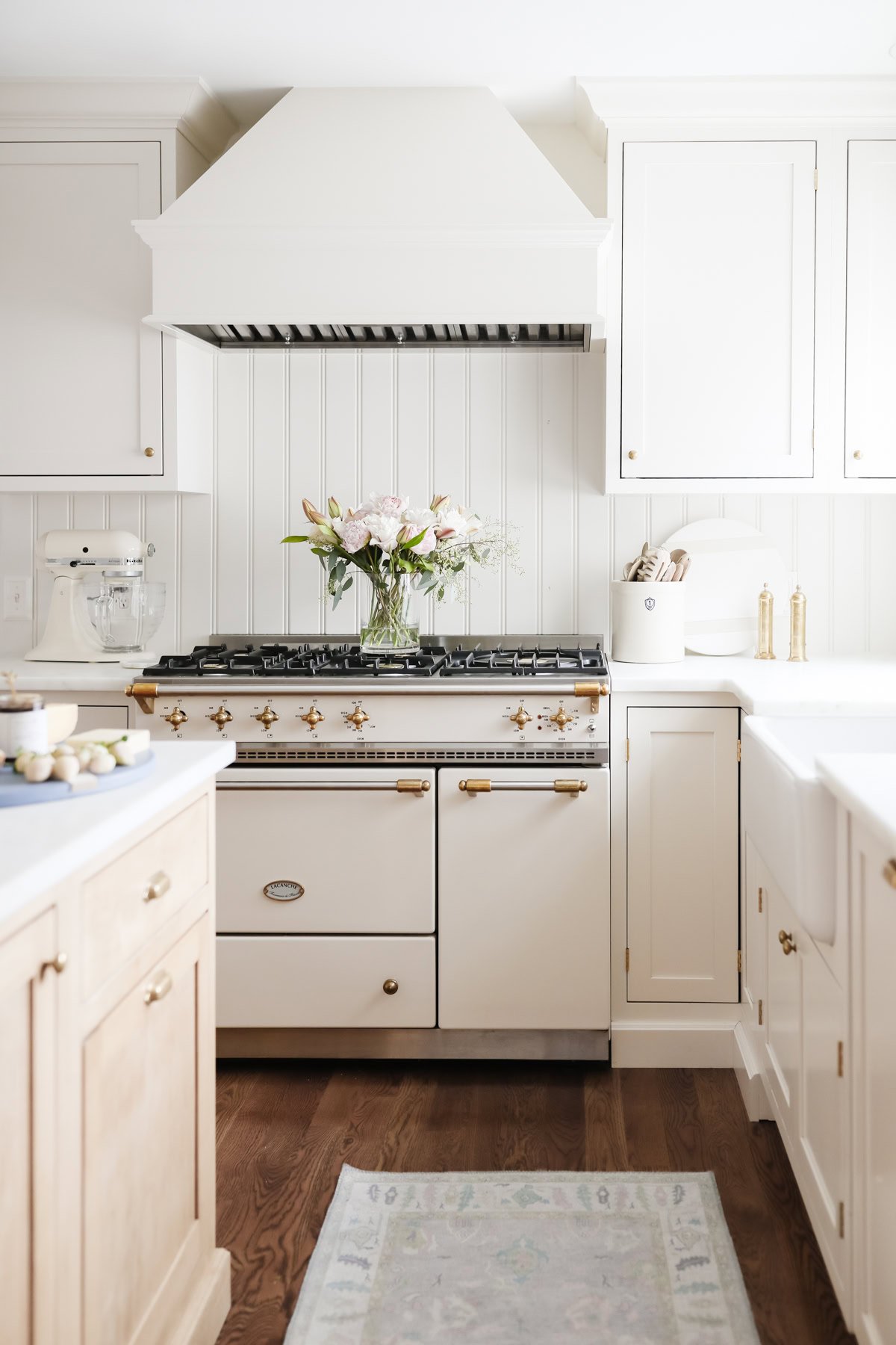 A white kitchen with a vintage-style stove, gold hardware, and decluttered white cabinets features wood floors and a vase of flowers on the stove.