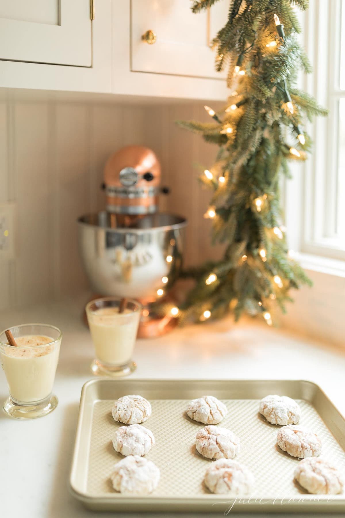 A white kitchen decorated with Christmas garland, and a cookie sheet full of eggnog Christmas cookies.