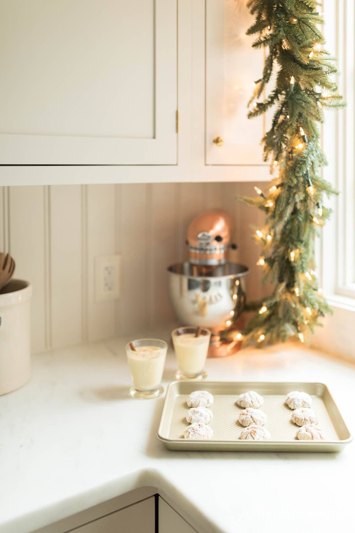 A white kitchen with garland over a window and Christmas cookies on a gold baking sheet. 