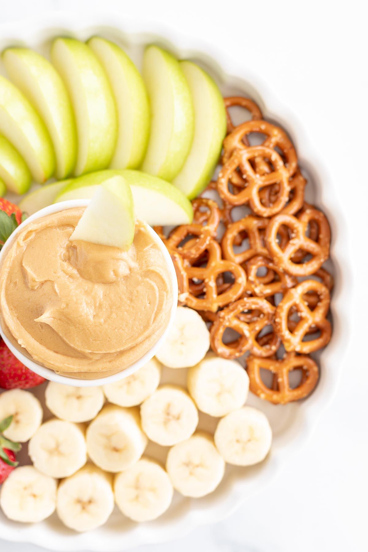 A peanut butter dip bowl in the center of fruit and pretzels. 