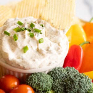 A hand dipping a ridged potato chip into a bowl of creamy dip garnished with chopped green onions, surrounded by broccoli, cherry tomatoes, and colorful bell peppers—try one of these best dip recipes for an easy snack.