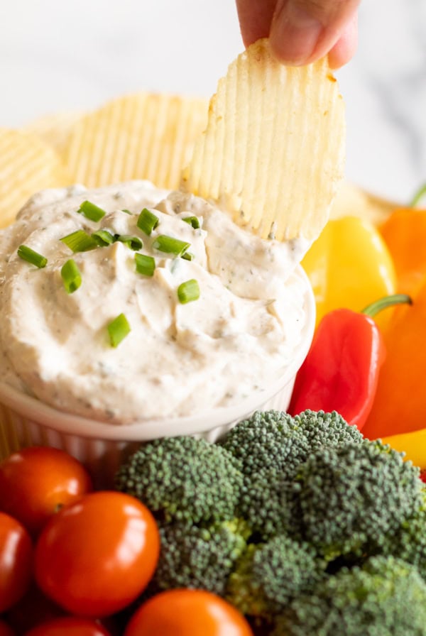 A hand dipping a ridged potato chip into a bowl of creamy dip garnished with chopped green onions, surrounded by broccoli, cherry tomatoes, and colorful bell peppers—try one of these best dip recipes for an easy snack.