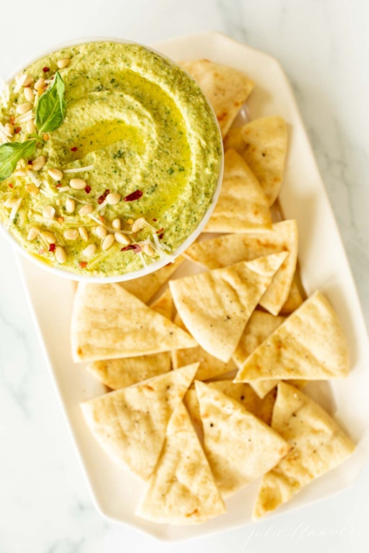 A white bowl full of basil pesto hummus on a platter full of pita triangles. 