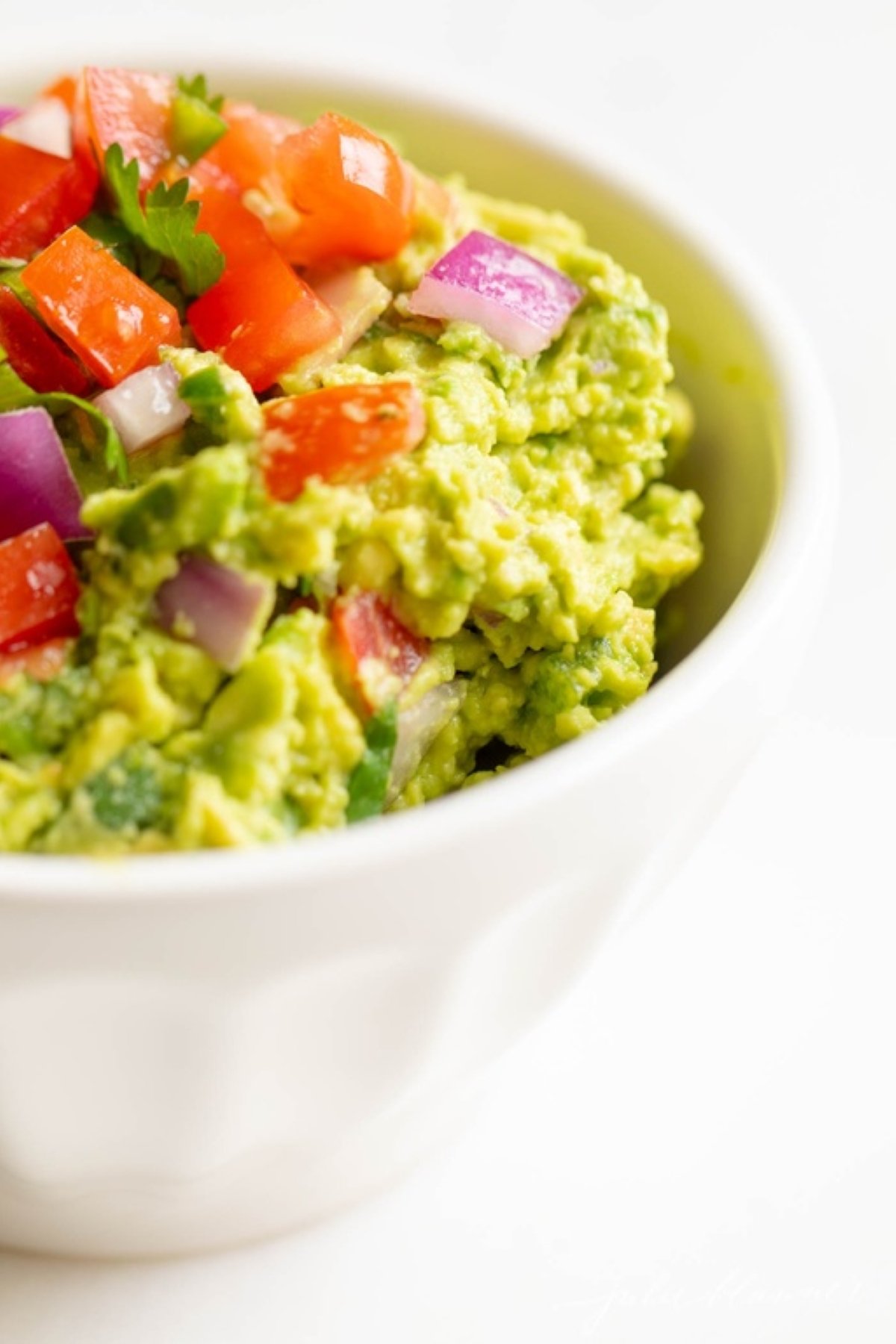 A white bowl on a white surface, filled with a homemade guacamole recipe.