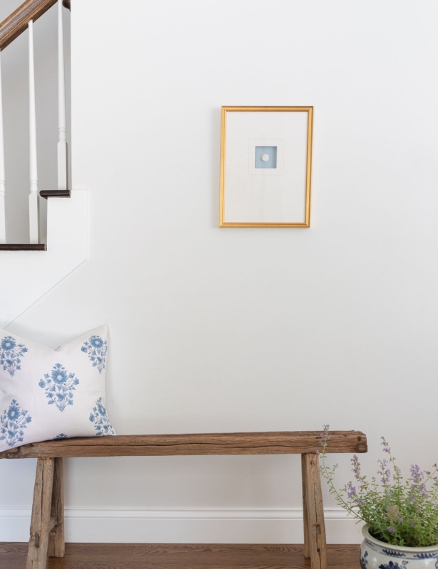 A gold framed intaglio in a white entryway of a home, wooden bench below.