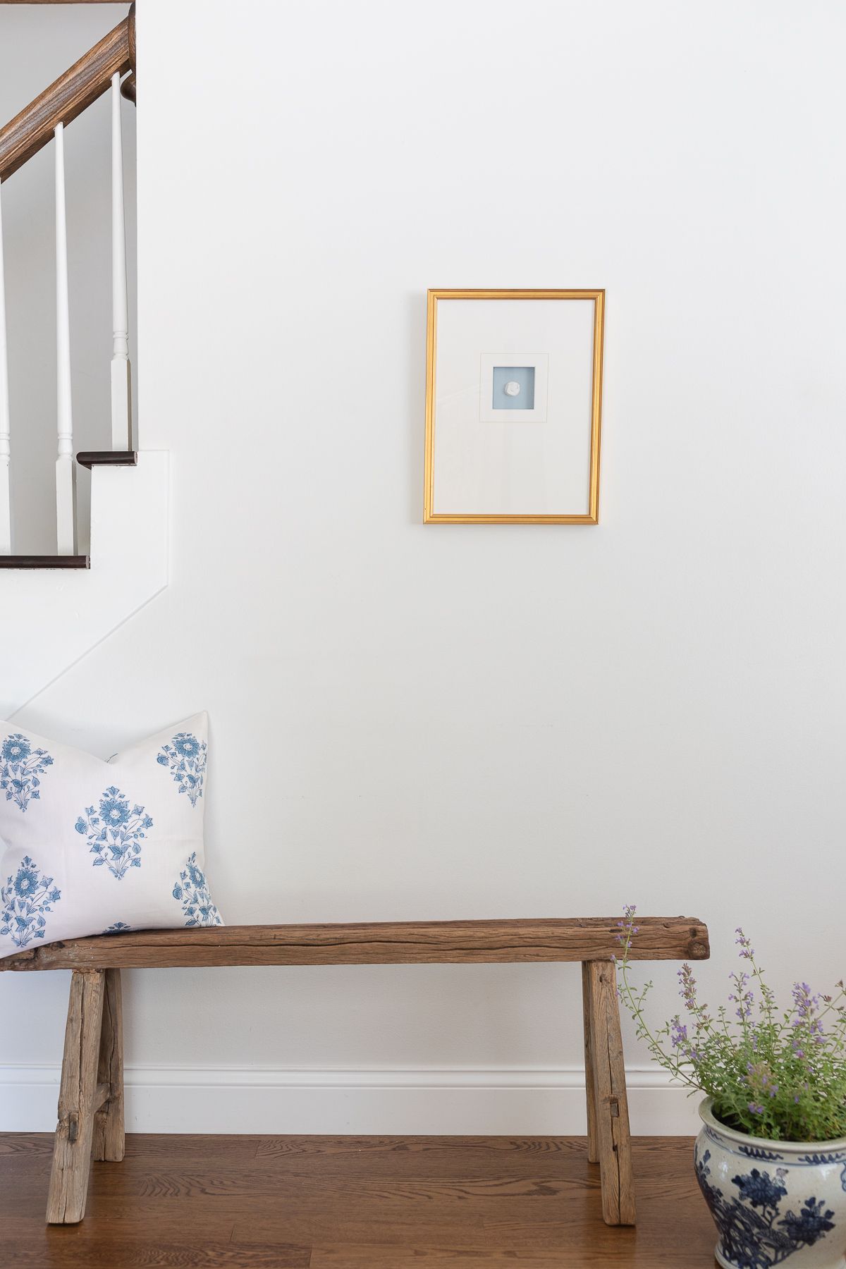 A gold framed intaglio in a white entryway of a home, wooden bench below. 