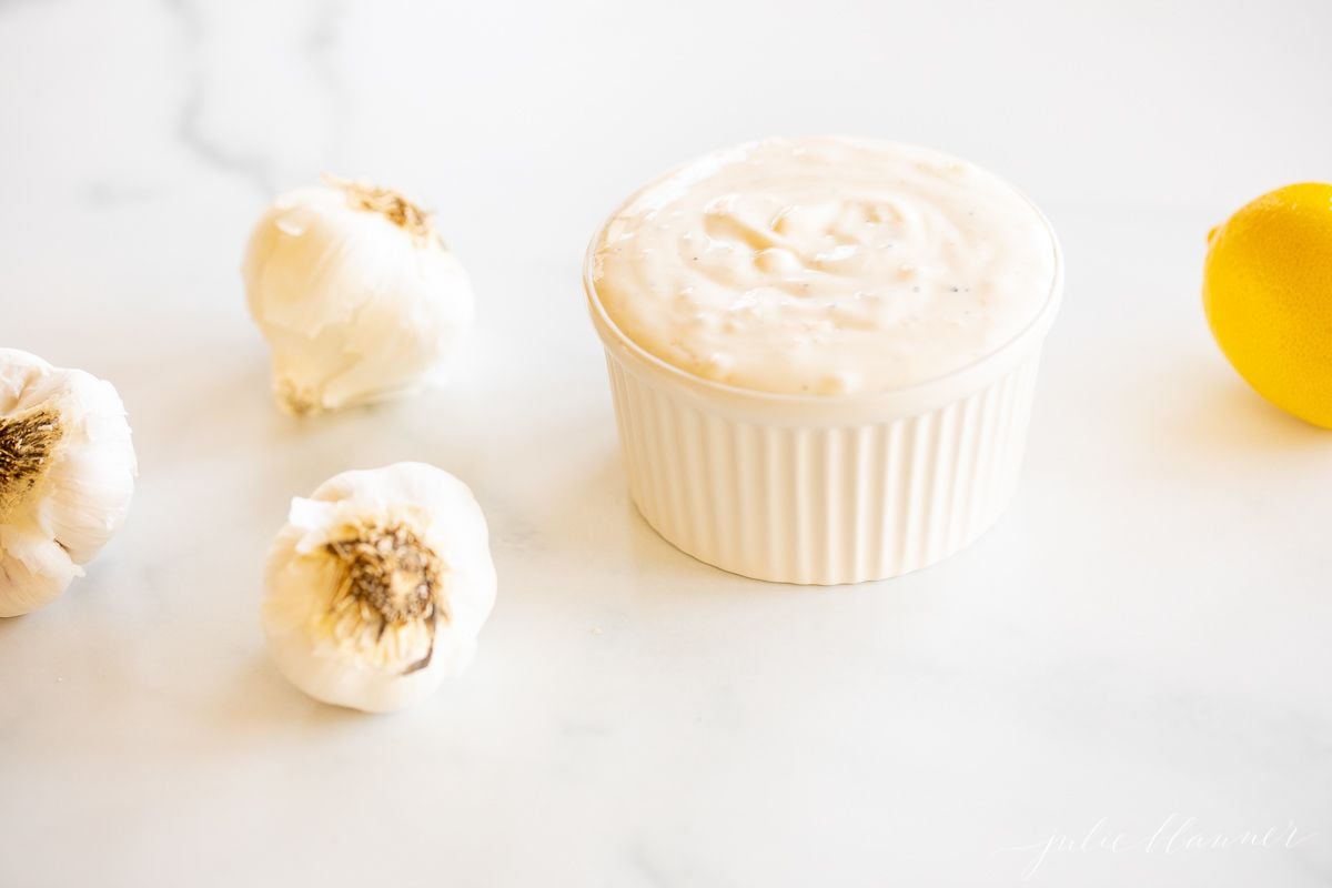 On a marble surface, a white ramekin dish full of fresh aioli, with heads of garlic to the side.