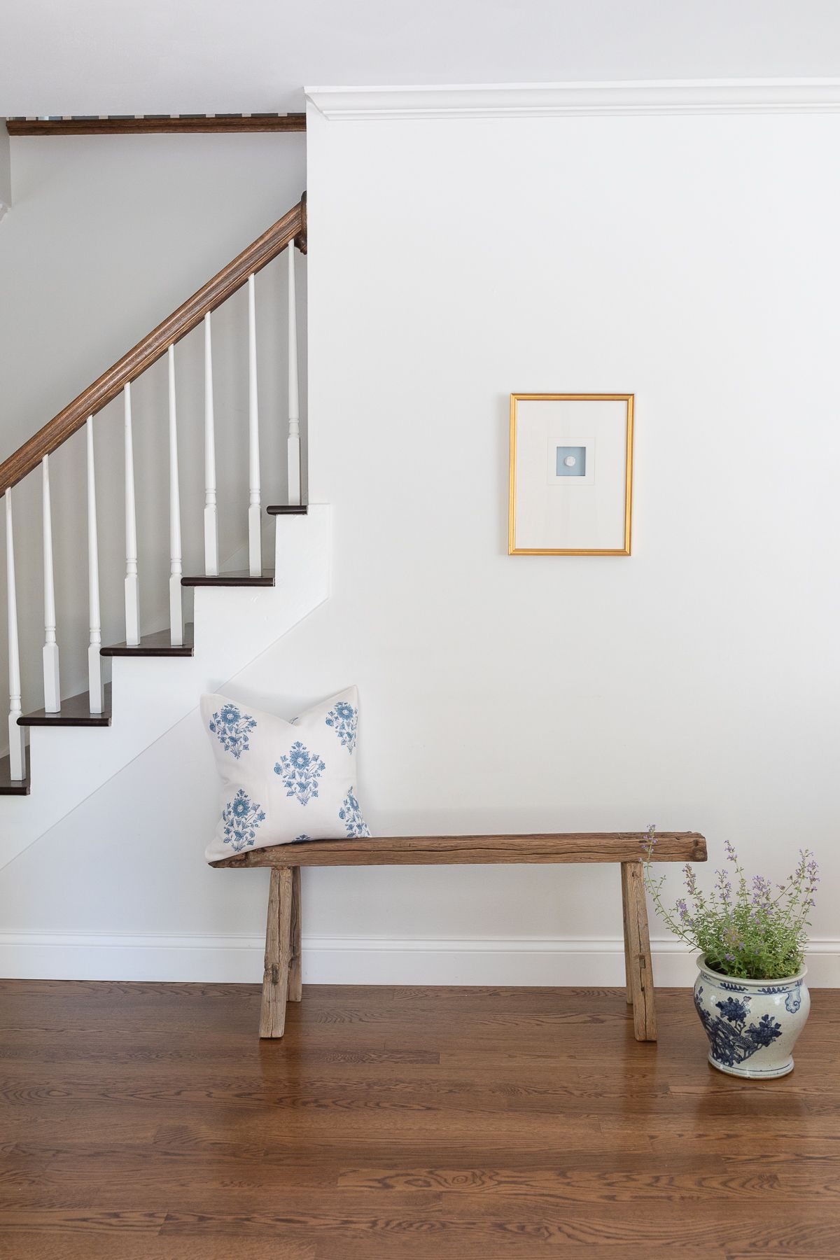 A gold framed intaglio in a white entryway of a home, wooden bench below. 