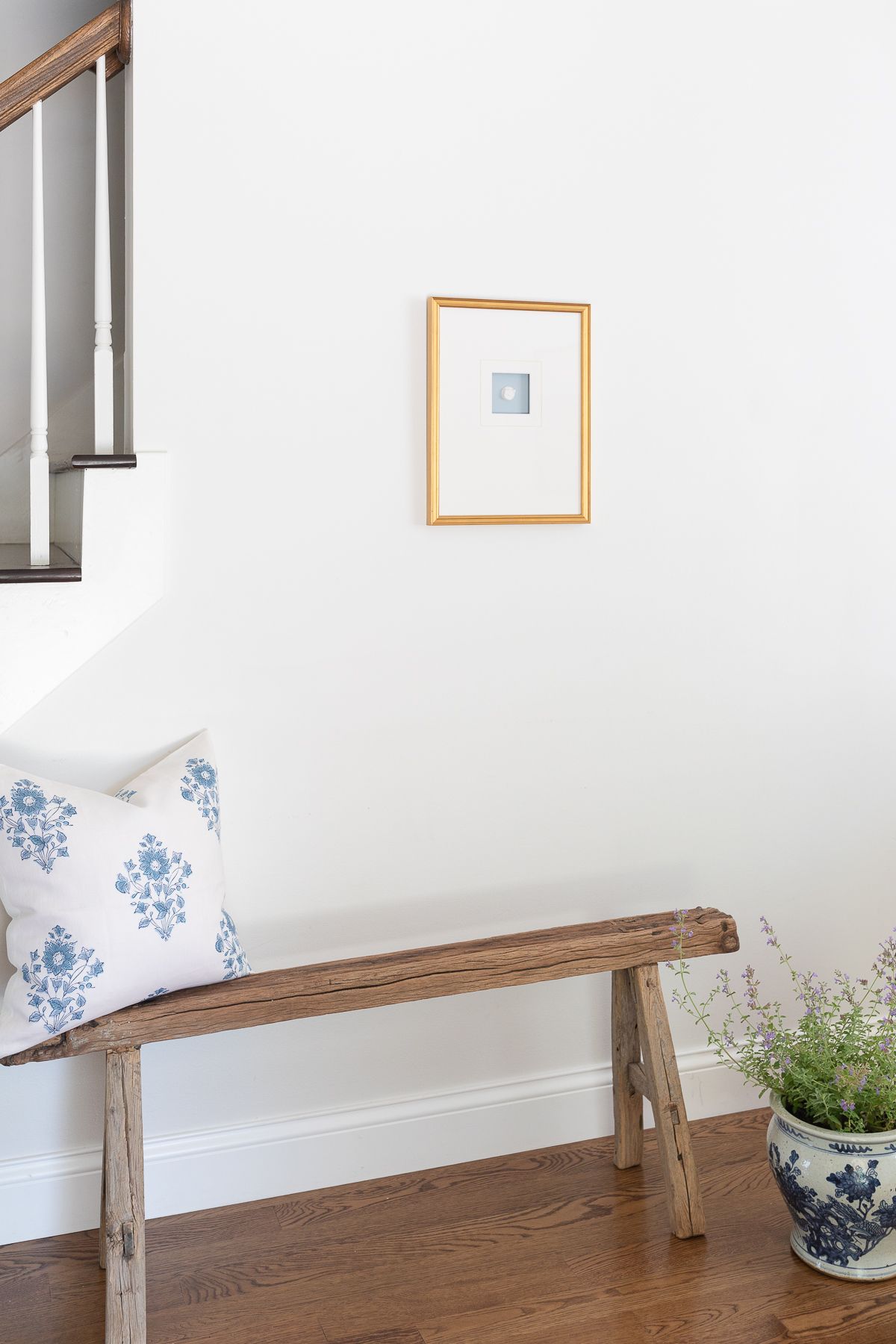 A gold framed intaglio in a white entryway of a home, wooden bench below.