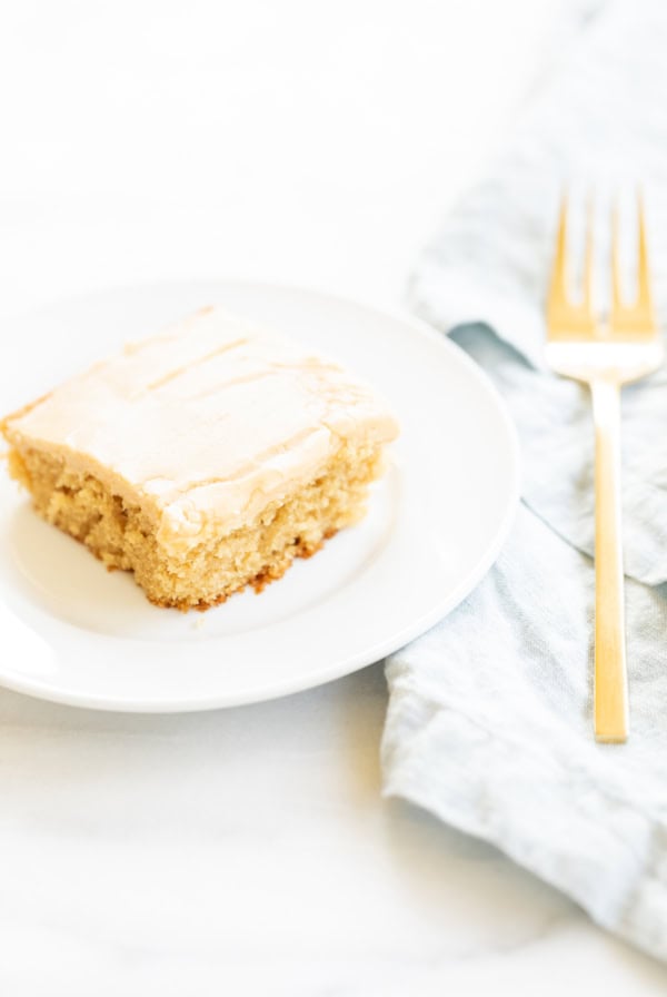 A piece of peanut butter frosted cake on a white plate with a gold fork on a light blue napkin, set against a bright background.
