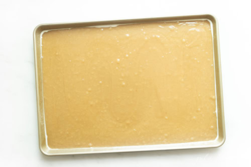 Unbaked peanut butter cake batter evenly spread in a rectangular metal baking tray, covered with a layer of plastic wrap.