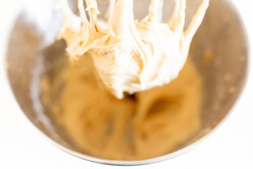 Close-up view of a mixing bowl with creamy peanut butter cake batter being lifted by mixing beaters, with a light background.