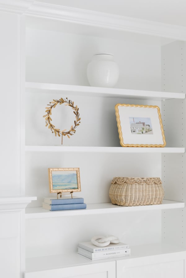 A white bookcase with a gold, scalloped frame and a wicker basket.