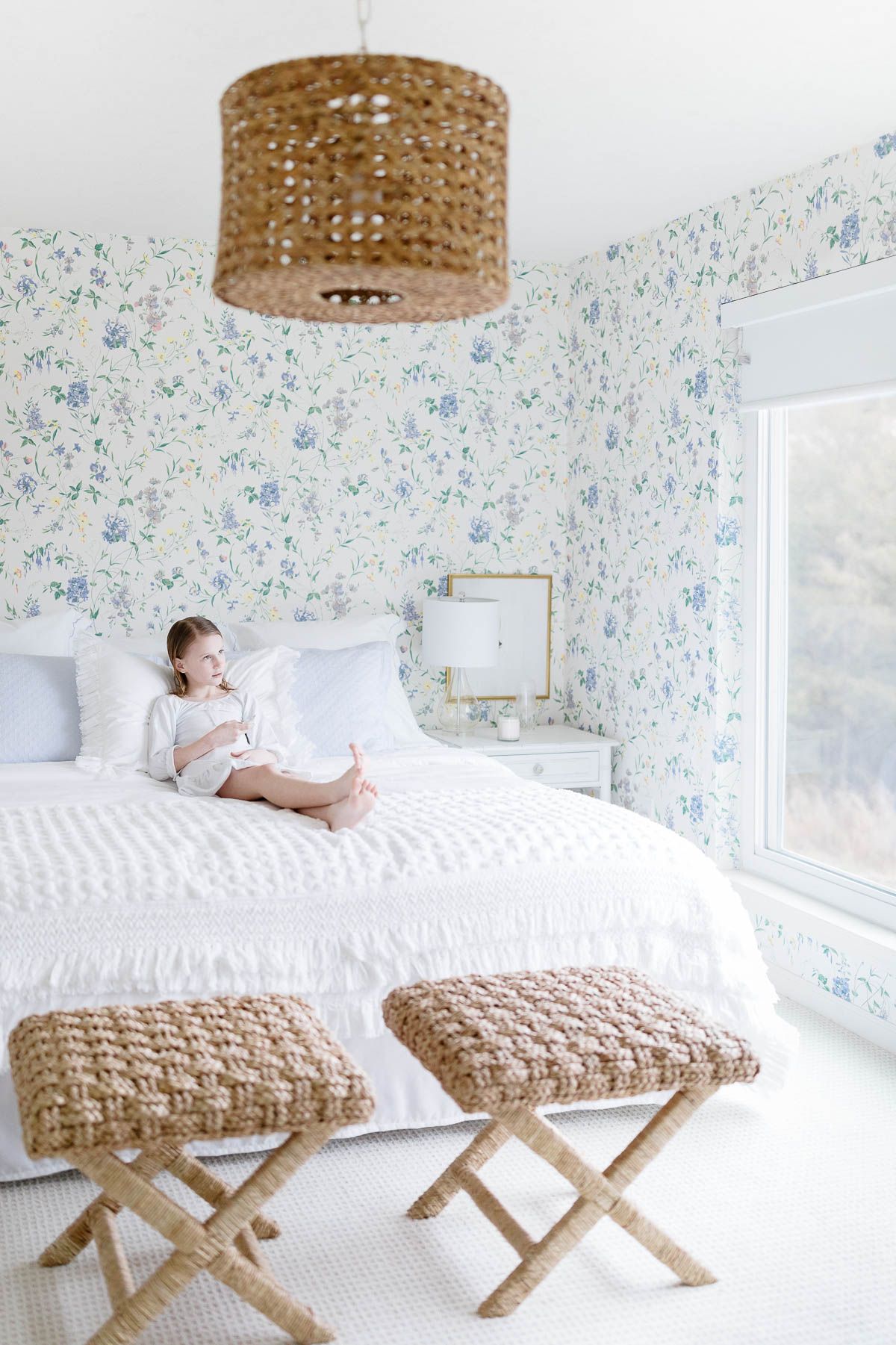 A little girl laying on a bed in a floral wallpapered bedroom, operating a motorized window shade. 