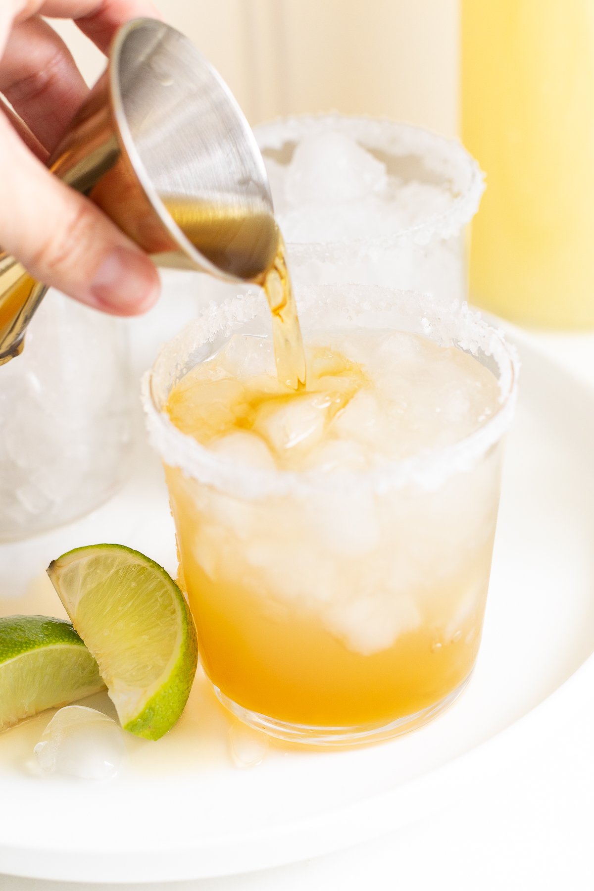 Pouring a golden-colored Cadillac margarita into a salt-rimmed glass with ice and lime wedges nearby.