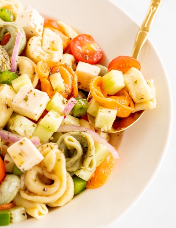 A white serving bowl full of tortellini pasta salad, gold serving spoon on the side.