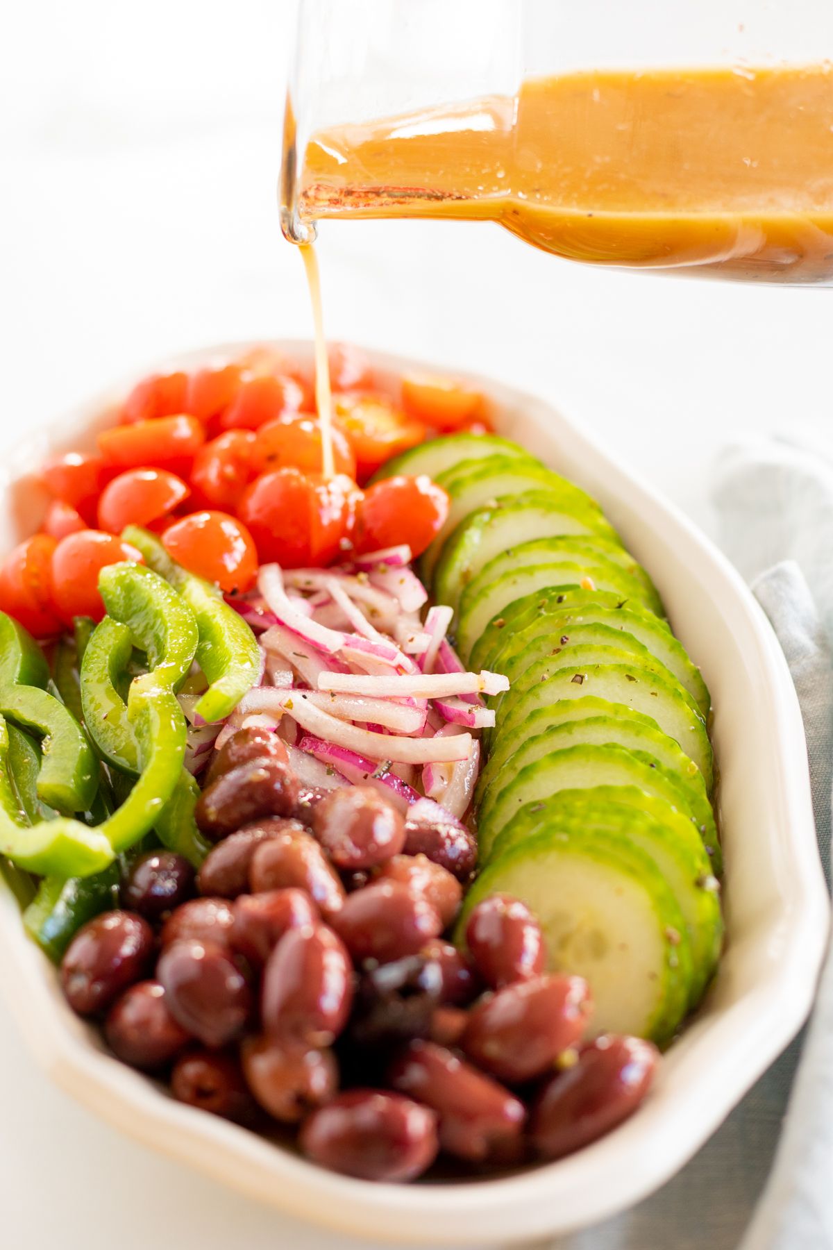 A small glass jar of Greek salad dressing pouring over a colorful Greek Salad on a white platter.