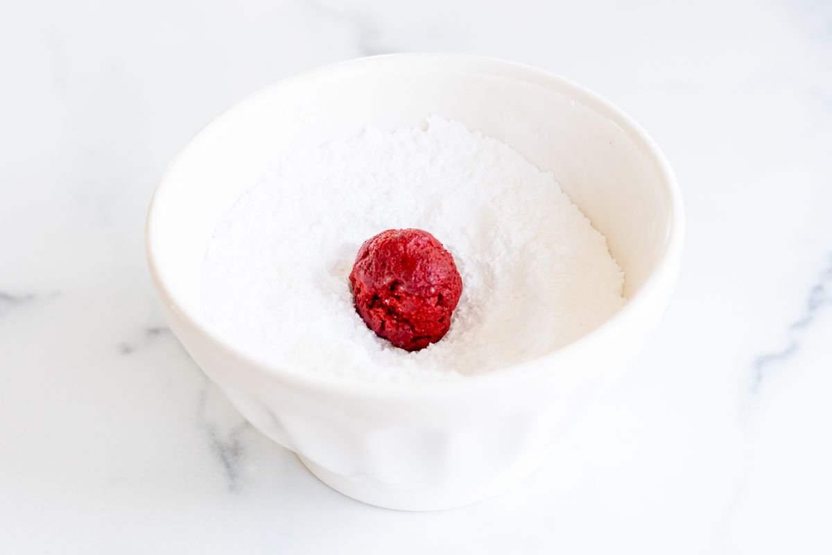 A red velvet gooey butter cookie in a bowl of powdered sugar.