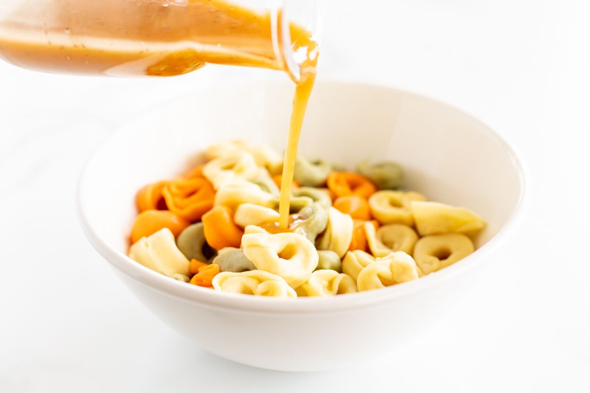 A white bowl filled with cooked tortellini pasta, and a bottle of pasta salad dressing being poured over the top. 