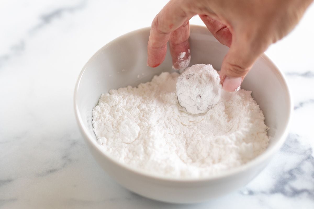 A hand dipping a red velvet gooey butter cookie into a bowl of powdered sugar.