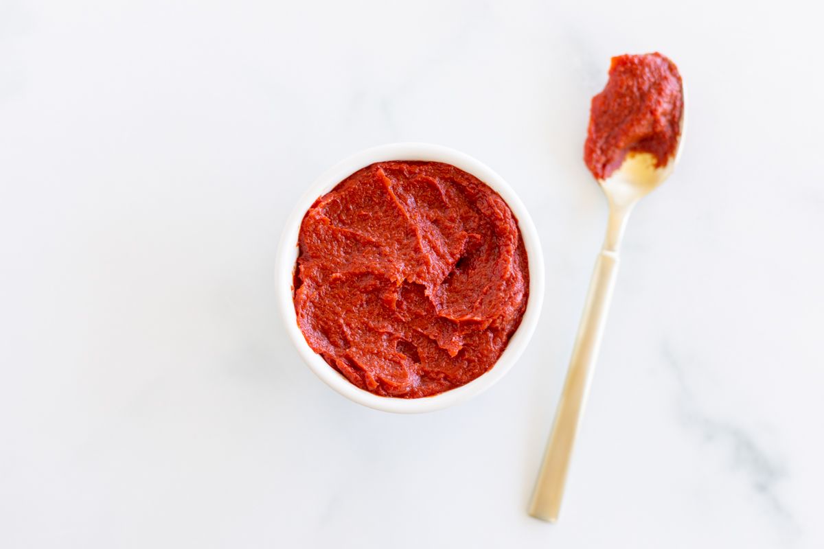 A small bowl of tomato paste on a marble countertop, gold spoon full of tomato paste substitute to the side.