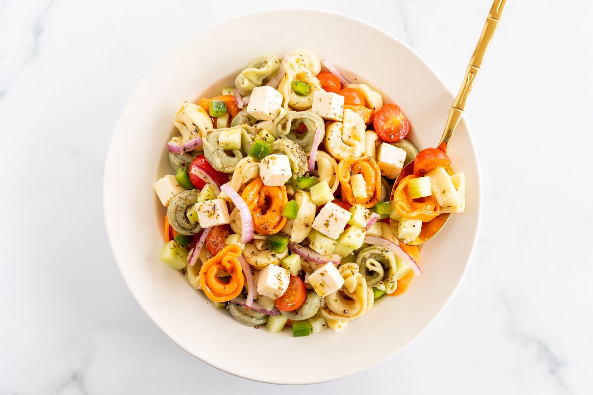 A white serving bowl full of tortellini pasta salad, gold serving spoon on the side.