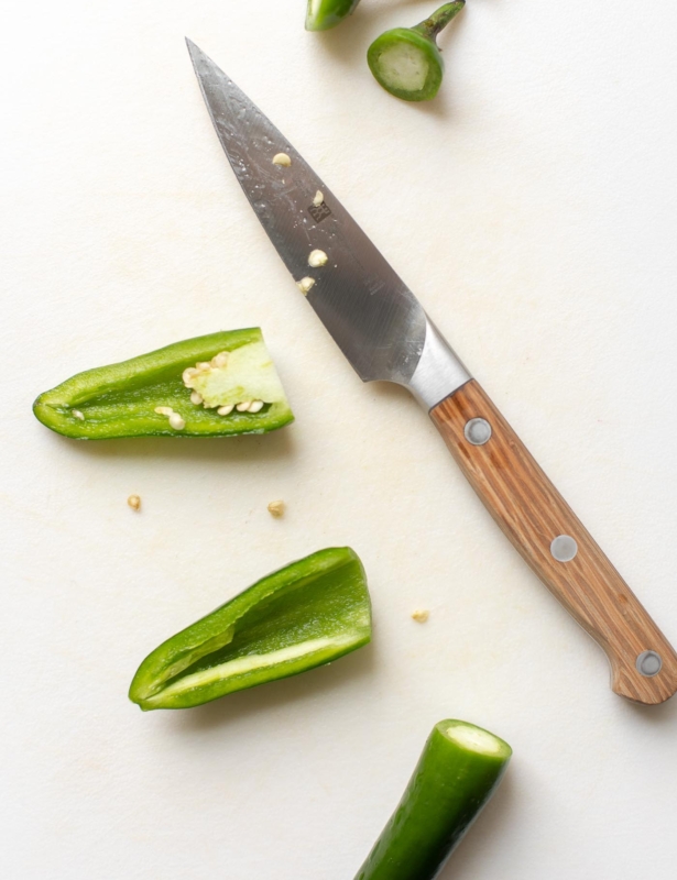 Overhead view of jalapeno peppers on cutting board with paring knife; one pepper has top cut off, the other is cut in half lengthwise