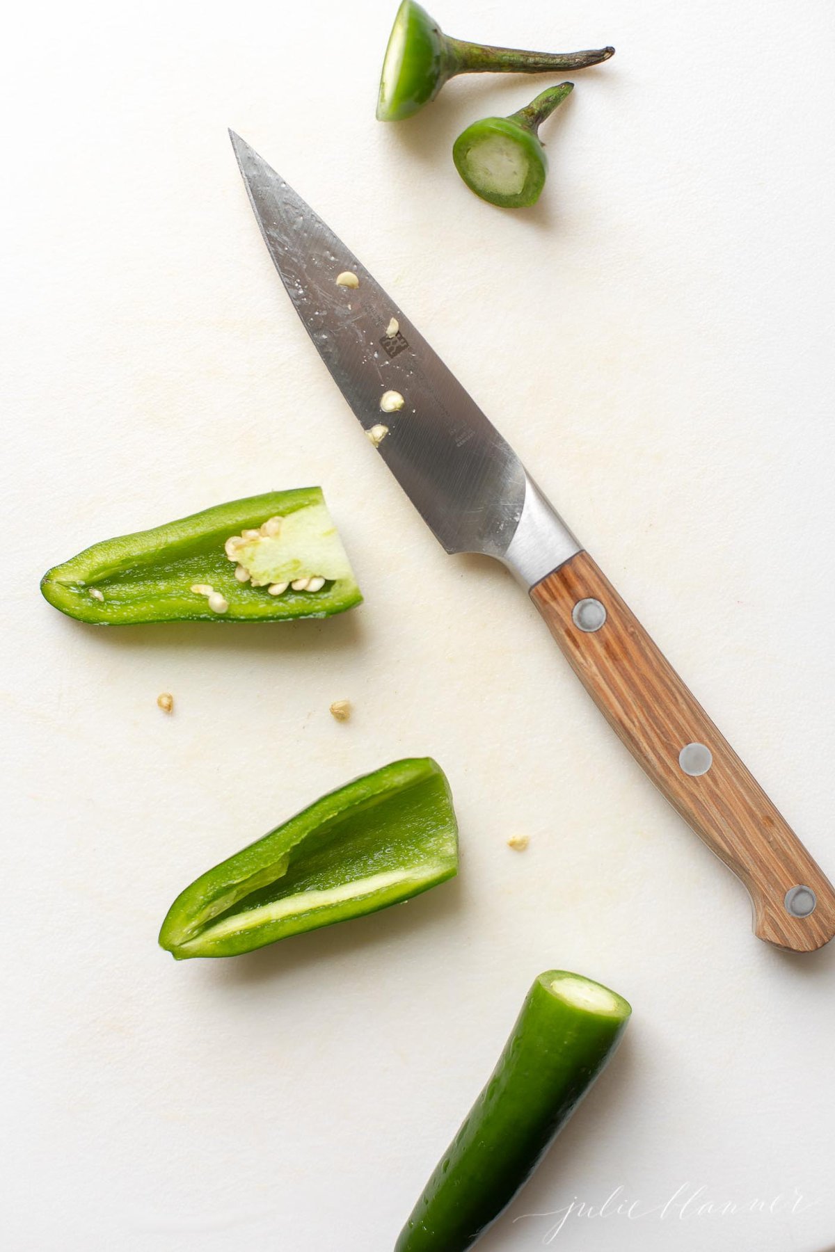 Overhead view of jalapeno peppers on cutting board with paring knife; one pepper has top cut off, the other is cut in half lengthwise