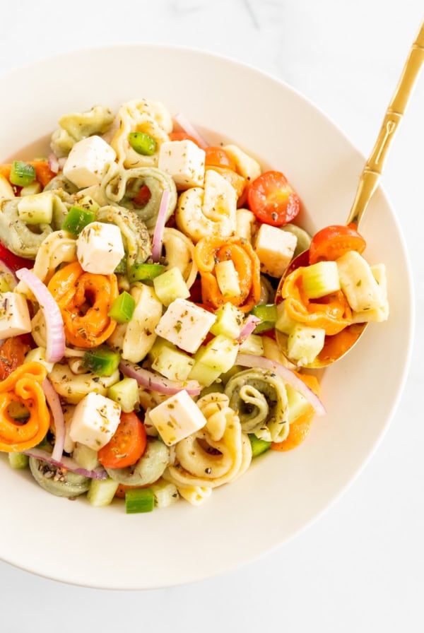 A colorful pasta salad with cherry tomatoes, cucumbers, red onions, and diced cheese, served in a white bowl with a gold fork.