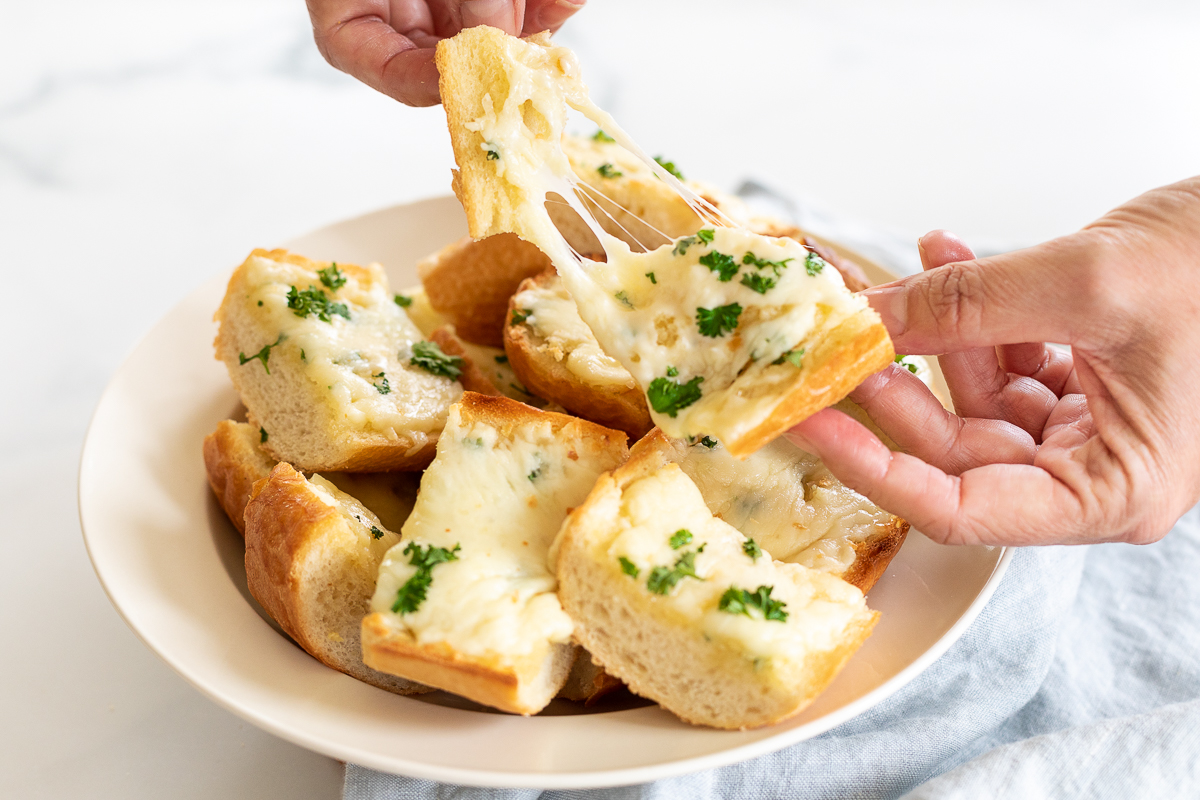 Garlic cheese bread in serving bowl, with hands pulling a piece apart to show cheese pull