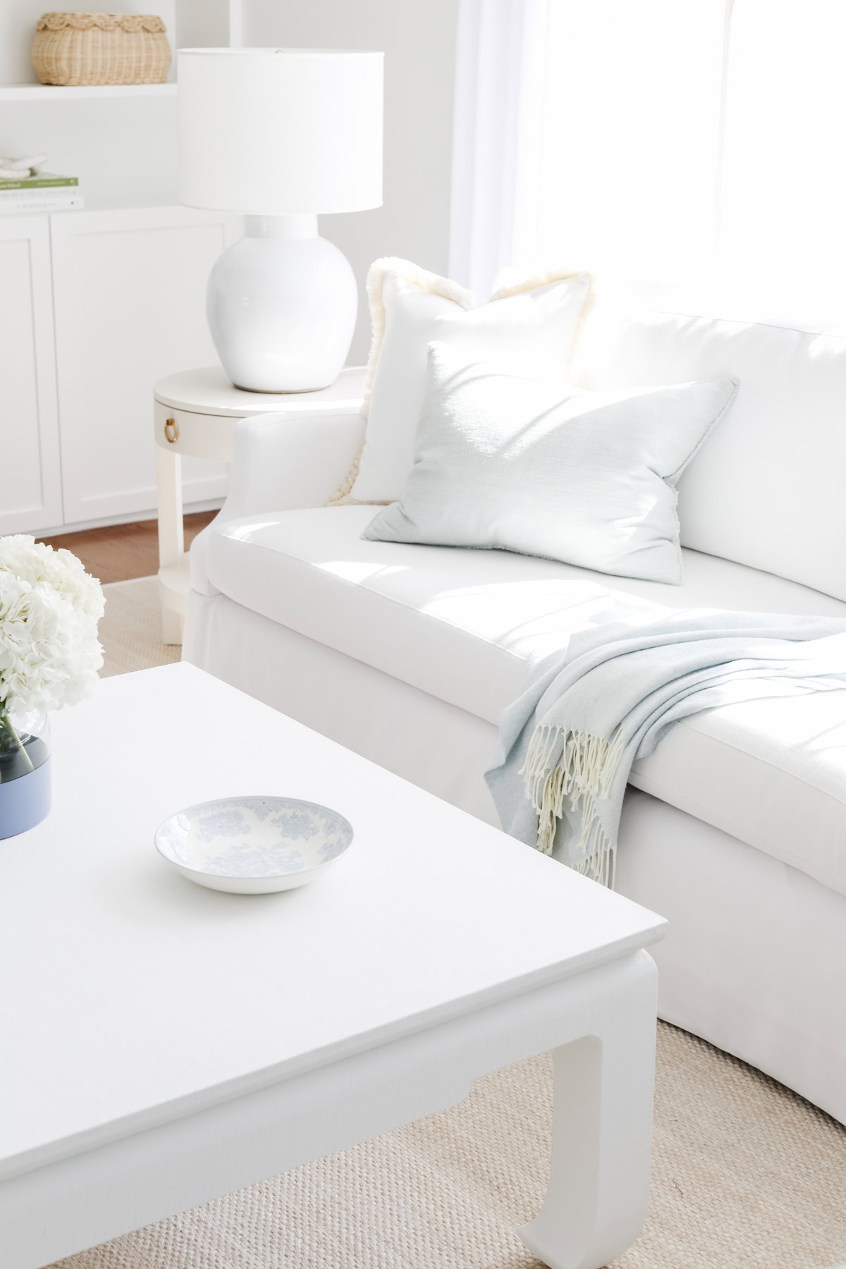 A white living room with a white sofa, light blue pillow, blanket, side table with a lamp, and a coffee table with a plate and vase of white flowers rests atop an elegant sisal rug with border.