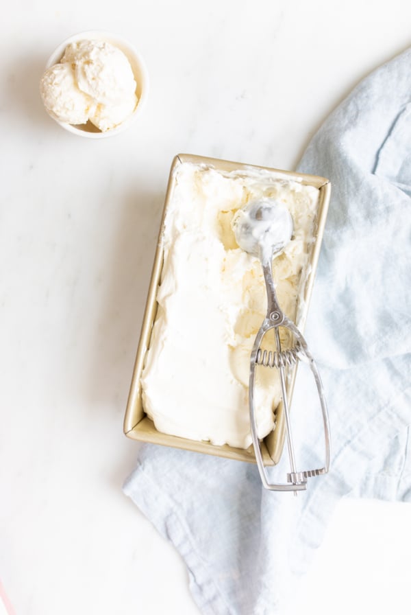 A container of no churn vanilla ice cream with a scoop and a bowl of scooped ice cream on a marble background.